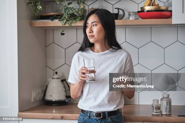 sad depressed asian woman holding pills and glass of water looking out window feeling upset while standing in kitchen at home, people coping with depression. mental health medications concept - pilule contraceptive photos et images de collection