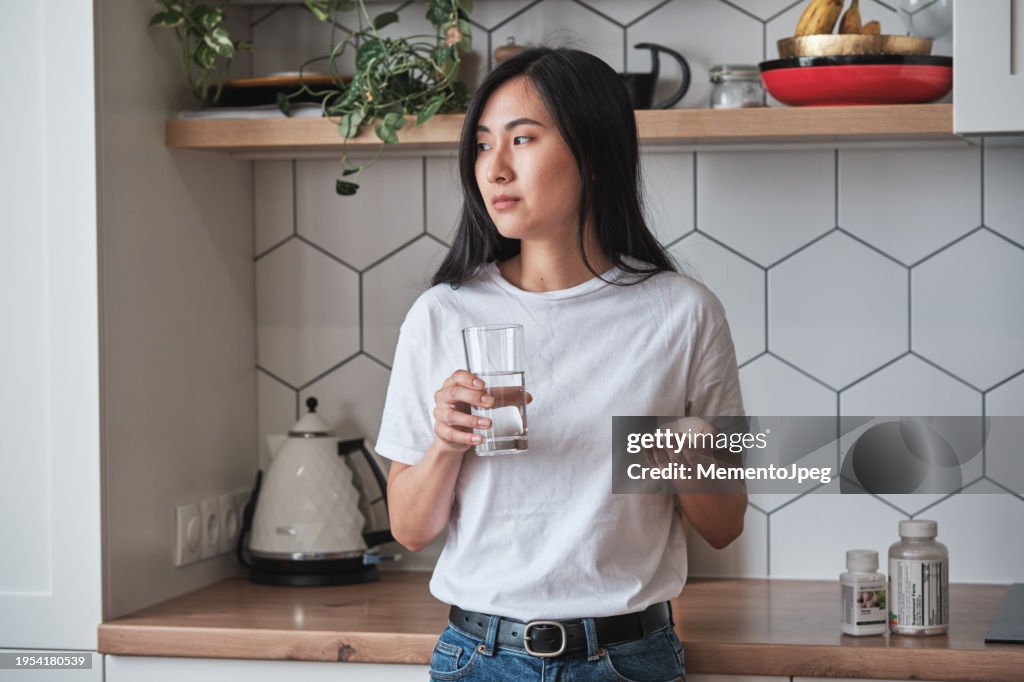 Sad depressed Asian woman holding pills and glass of water looking out window feeling upset while standing in kitchen at home, people coping with depression. Mental health medications concept