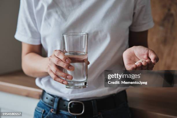 woman holding pills and glass of water while standing in kitchen at home - taking pills stock pictures, royalty-free photos & images