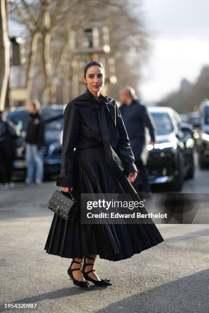 Bettina Looney wears earrings, a black shirt, black tie, pleated midi skirt, quilted leather bag, pointed kitten heels pointed pumps / shoes with bow...