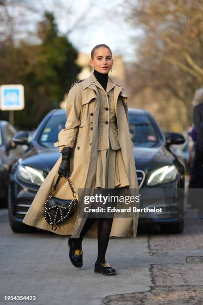 Caroline Daur wears earrings, a black turtleneck pullover, a beige long trench coat, black leather gloves, a leather Dior Saddle bag, a padded Dior...