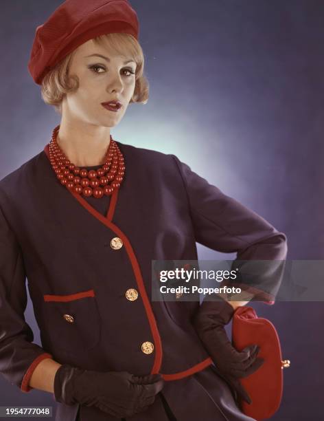 Posed studio portrait of a female fashion model wearing a navy blue jacket with red piping and metallic gold buttons over a matching blue pleated...