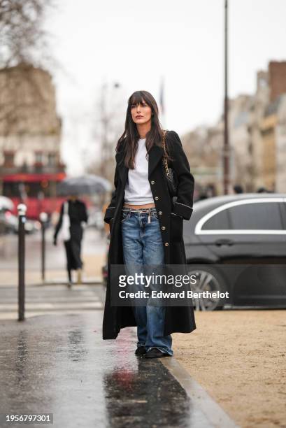 Leia Sfez wears a white t-shirt, a black quilted leather Chanel bag, a black long coat, a belt, blue flared denim jeans pants, shoes, outside Chanel,...