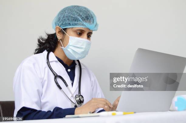one indian physician woman or lady or female doctor wearing surgical mask, cap, and white lab coat and a stethoscope around her neck, sitting and working focussed on a laptop  over grey horizontal background with copy space for text - lady grey background bildbanksfoton och bilder