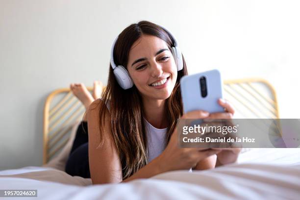 happy young woman listening to music with headphones app mobile phone app lying on bed. - badalona - fotografias e filmes do acervo