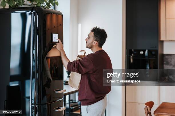 man effortlessly uses his smart fridge while holding a shopping bag - refrigerator stock pictures, royalty-free photos & images