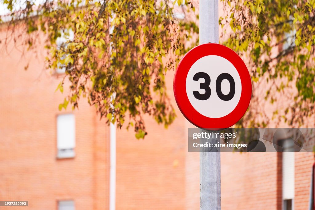 A circular traffic sign with the number thirty attached to a metal pole, branches of a tree and the wall of a building can be seen.
