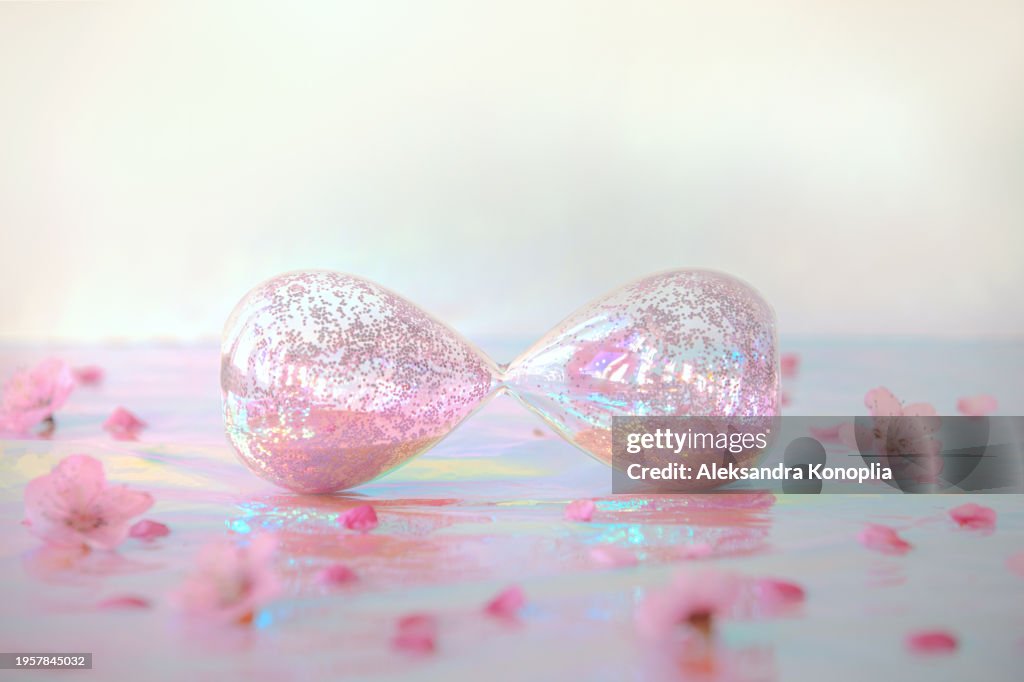Sparkling pink hourglass lying on holographic foil background with peach tree flowers spring bloom. Front view, copy space. Eternity concept