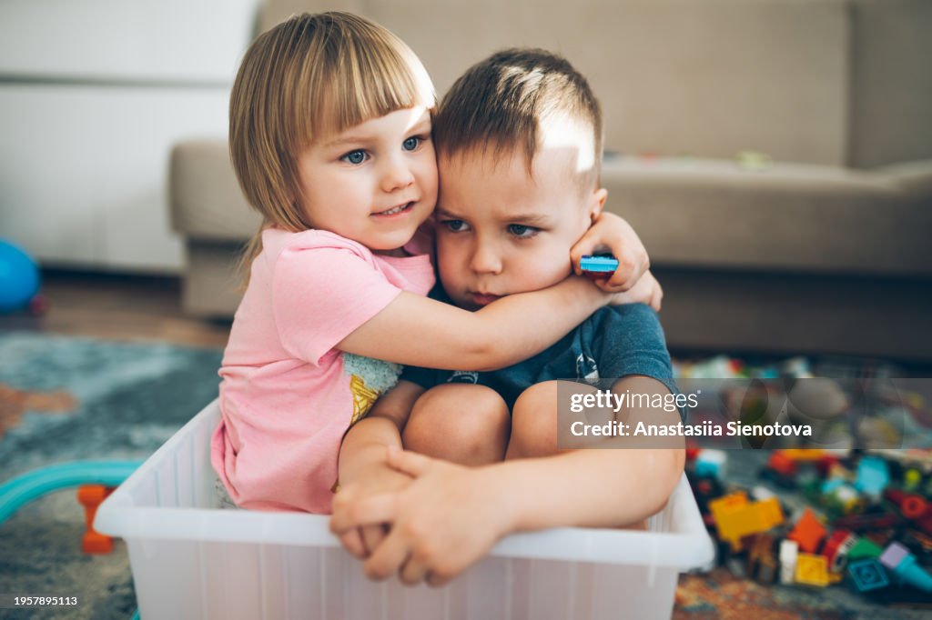 A girl hugging an upset boy at home among toys