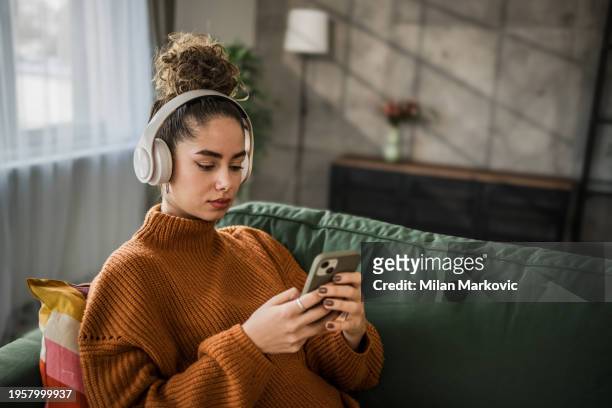 excited young woman listening to music on wireless headphones over a mobile app while sitting on a comfortable sofa - people podcasting stock pictures, royalty-free photos & images