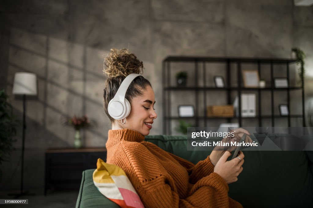 Giovane donna eccitata che ascolta musica su cuffie wireless tramite un'app mobile mentre è seduta su un comodo divano