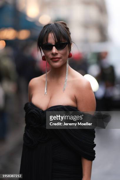 Fashion week guest is seen wearing a black off-the-shoulder dress with ruffles and roses, long rhinestone earrings with red roses, a small black...
