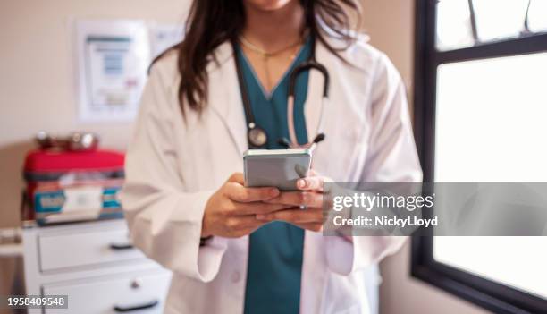 médecin utilisant un téléphone debout dans une salle d’examen d’hôpital - blouse dexamen médical photos et images de collection