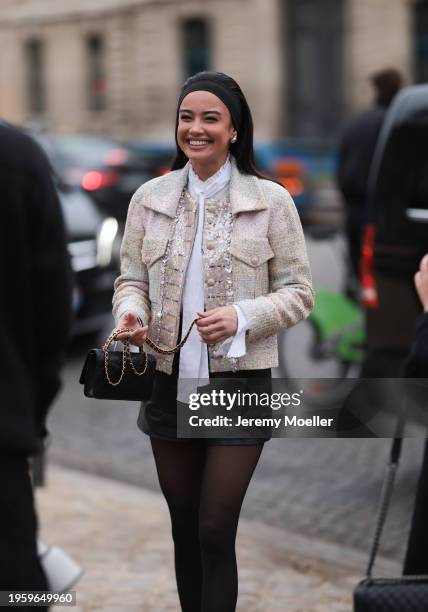 Kelsey Merritt is seen wearing a black hairband, golden creoles, a white blouse with a long bow, a beige tweed jacket with pearls, black leather...