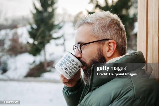side view of man standing on cabins patio, drinking hot tea, coffee and enjoying beautiful winter day. handsome man spending relaxing, stress-free winter weekend in cabin in mountains, enjoying alone time. - hygge stockfoto's en -beelden