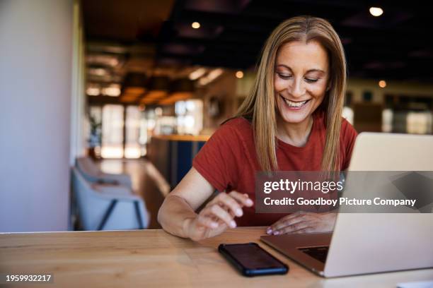 mature businesswoman smiling at a phone text while working on a laptop - file sharing stock pictures, royalty-free photos & images