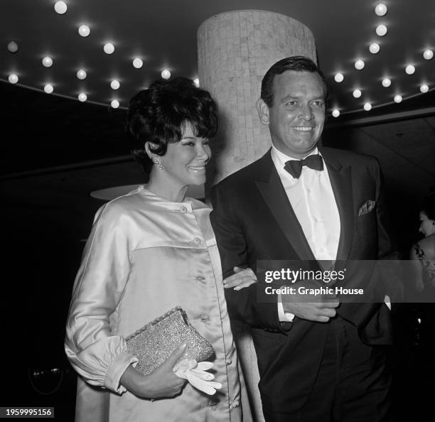 American fashion model Ellie Graham, wearing a button-front satin coat, arm-in-arm with her husband, David Janssen, who wears a tuxedo and bow tie,...