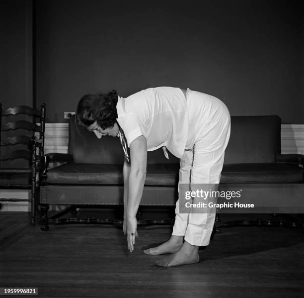 Woman wearing a white short-sleeved shirt and white trousers bends over, her fingertips almost touching the floor, as she exercises barefoot, with a...