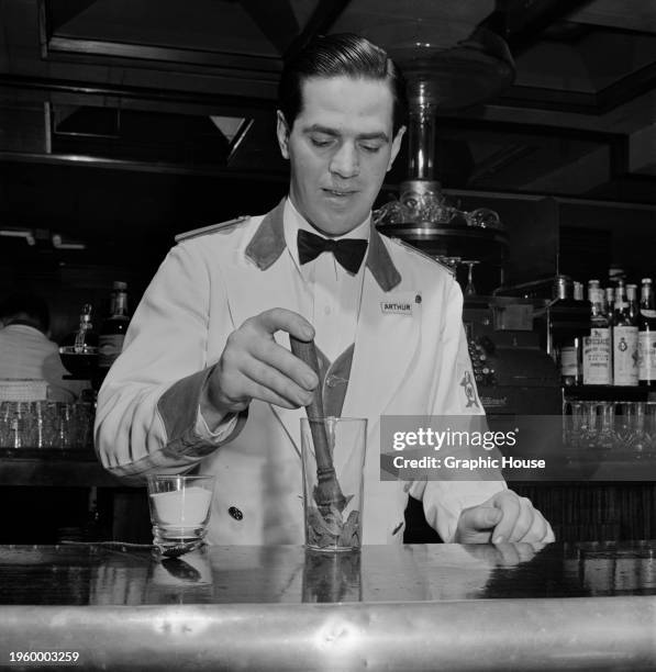 Bartender, with 'Arthur' on his badge, preparing a cocktail, using a muddler to press mint leaves in a glass, behind the bar at a party, United...
