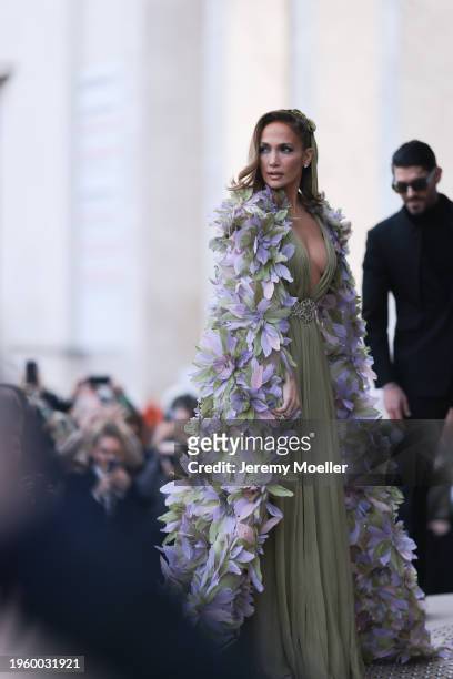 Jennifer Lopez is seen wearing an olive green dress with V-neck and silver belt, a feather cape with purple, green feathers and an olive green velvet...