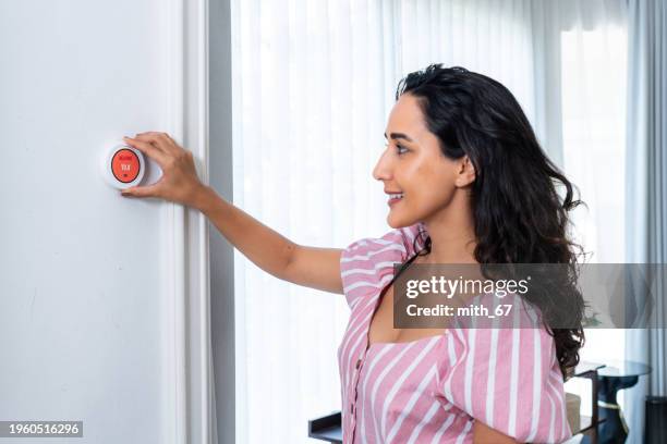 una bella donna adulta che regola la temperatura su un termostato intelligente sul muro del soggiorno di casa durante l'inverno - termostato foto e immagini stock