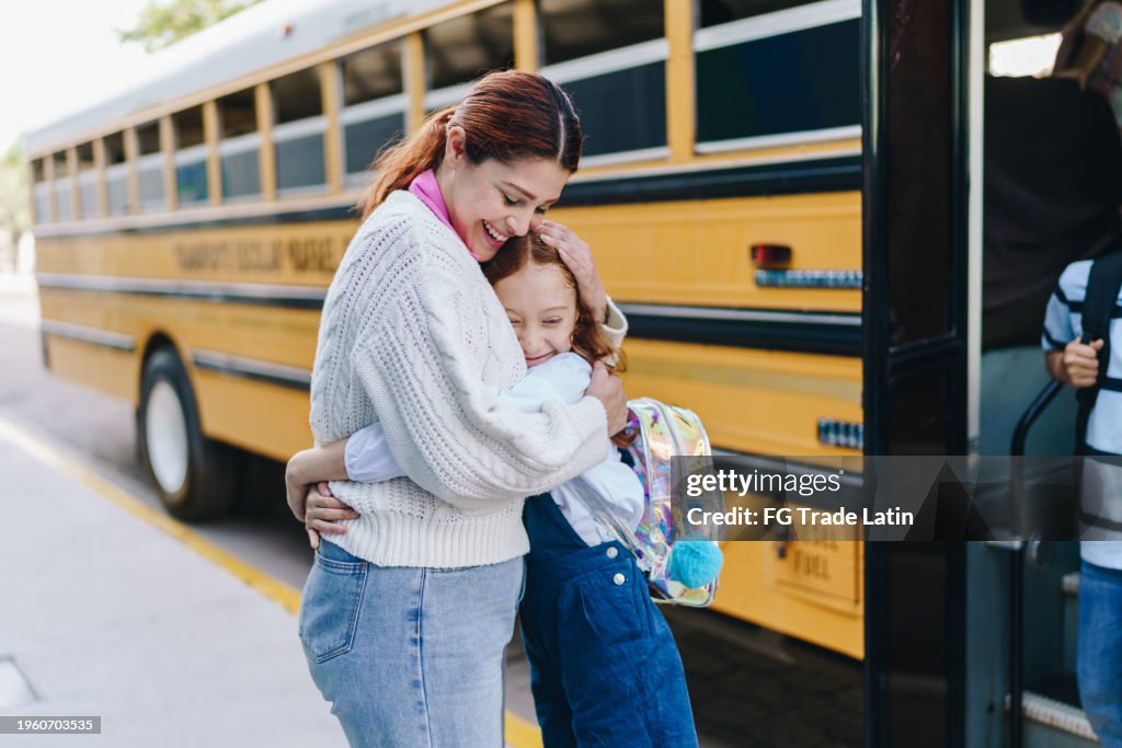 Child student girl embracing mother before getting on the school bus on the street