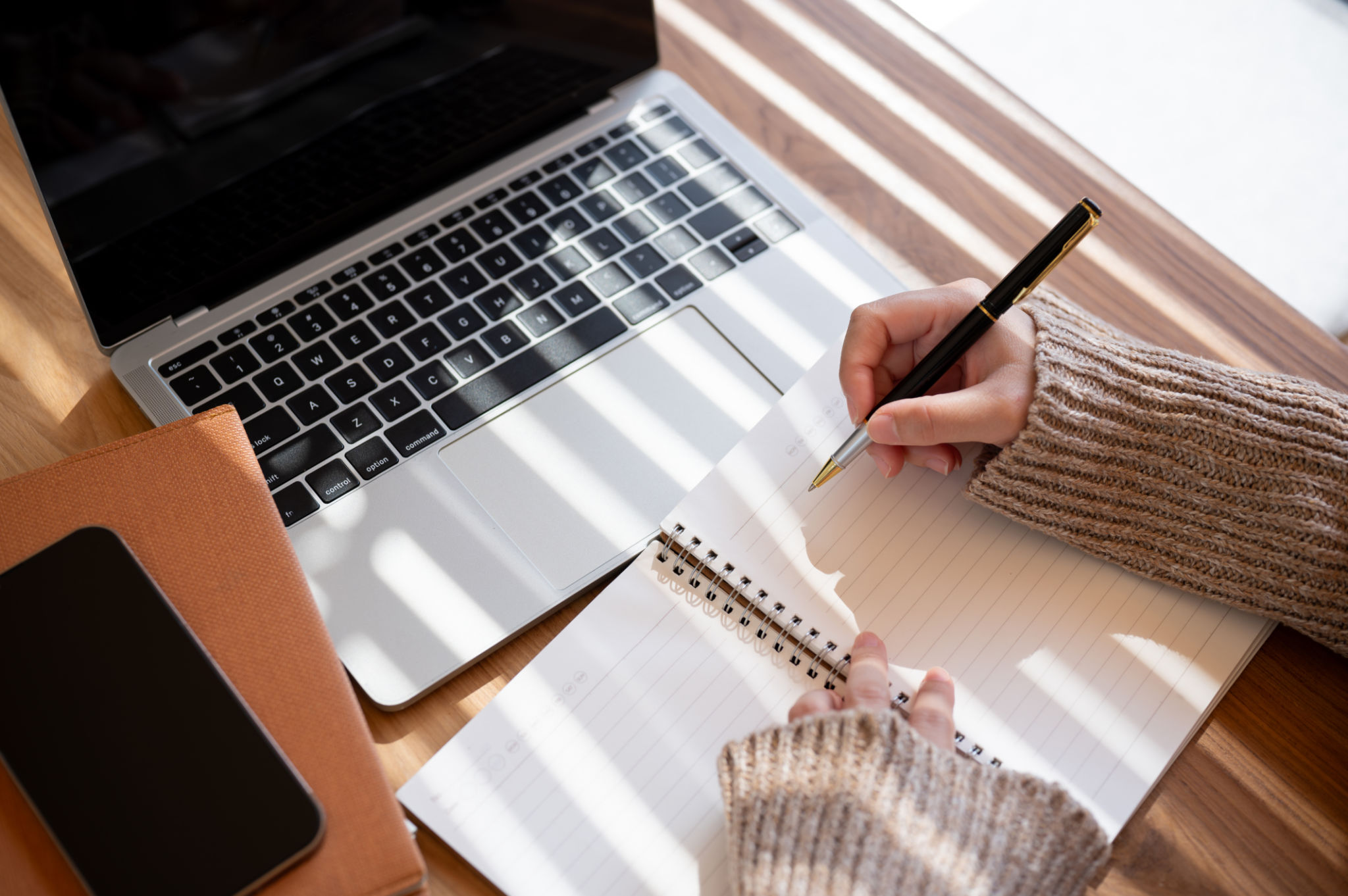 A top-view image of a woman is taking notes in her notebook while working on her laptop indoors. A top-view image of a woman is taking notes in her notebook while working on her laptop indoors.