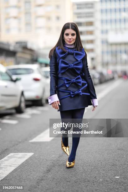 Gessica Kayane aka Gkay wears earrings, a purple turtleneck top, a navy blue mini dress with gathered sleeves and oversized blue bow details in...