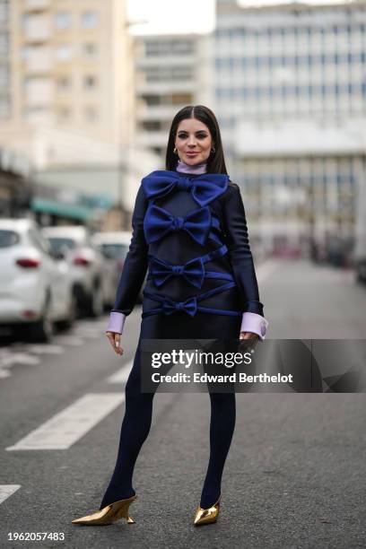 Gessica Kayane aka Gkay wears earrings, a purple turtleneck top, a navy blue mini dress with gathered sleeves and oversized blue bow details in...