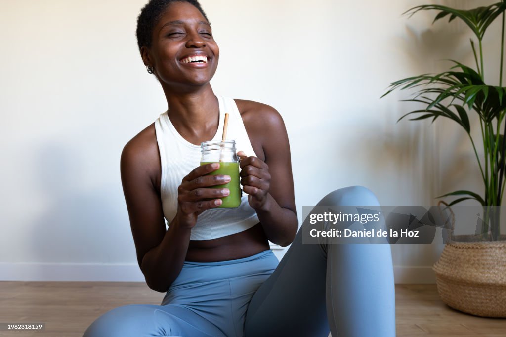 Happy young African American woman wearing fitness clothes drinking healthy green juice after yoga at home laughing. Happy Black female enjoying green smoothie after workout.