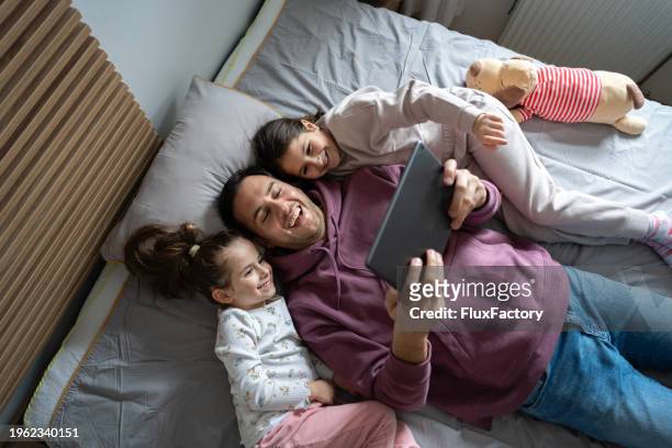 père regardant un dessin animé sur une tablette numérique tout en étant allongé dans le lit avec ses filles et en riant - movie film reel photos et images de collection