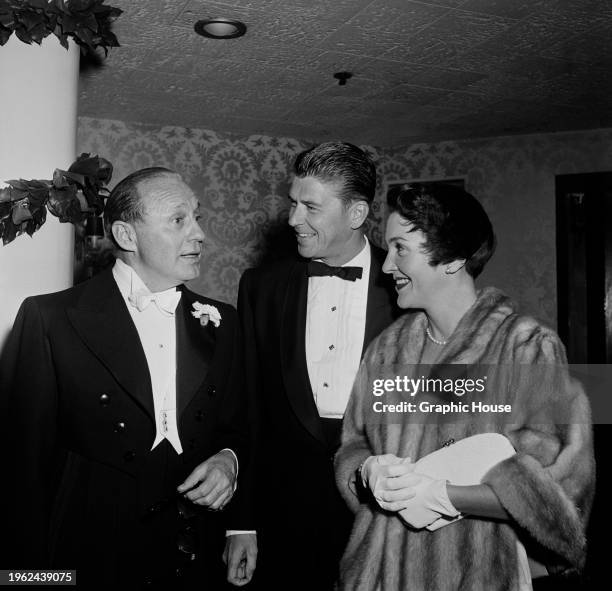 American comedian Jack Benny, wearing a black suit over a white shirt and white bow tie, in conversation with American actor Ronald Reagan, who wears...