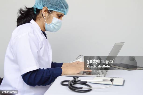one indian physician woman or lady or female doctor wearing surgical mask, cap, and white lab coat and a stethoscope around her neck, sitting and engrossed working on a laptop  over grey horizontal background - lady grey background bildbanksfoton och bilder