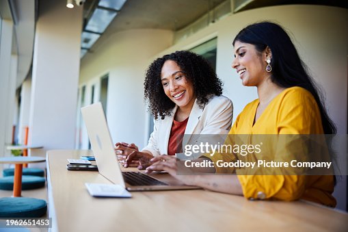 Two businesswomen smiling and working on a laptop at an office table Two businesswomen smiling and working on a laptop at an office table