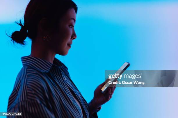 side view of young asian woman in silhouette using smartphone against illuminated blue screen - shopping online blue stock pictures, royalty-free photos & images