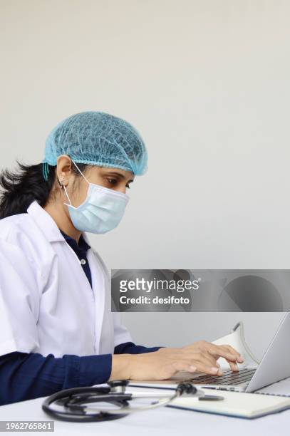 one indian physician woman or lady or female doctor wearing protective mask, cap, and white lab coat, sitting and working focussed on a laptop  over grey vertical background with stethoscope on table and copy space for text - lady grey background bildbanksfoton och bilder