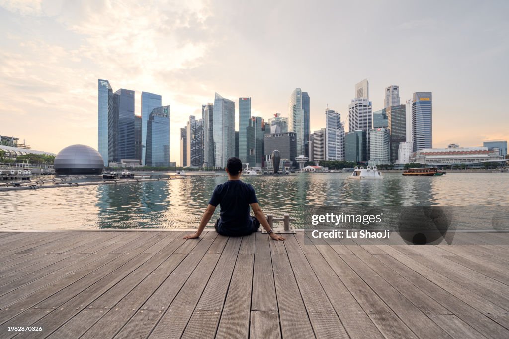 The young man sat on the dock with urban skyline and skyscrapers in Marina Bay Singapore.