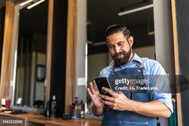 barber using mobile phone at barber shop - mexican men - fotografias e filmes do acervo