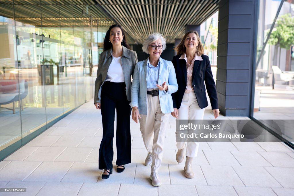 Full length photo of three empowered mature businesswomen walk purposefully into the office, looking cheerfully at the camera.