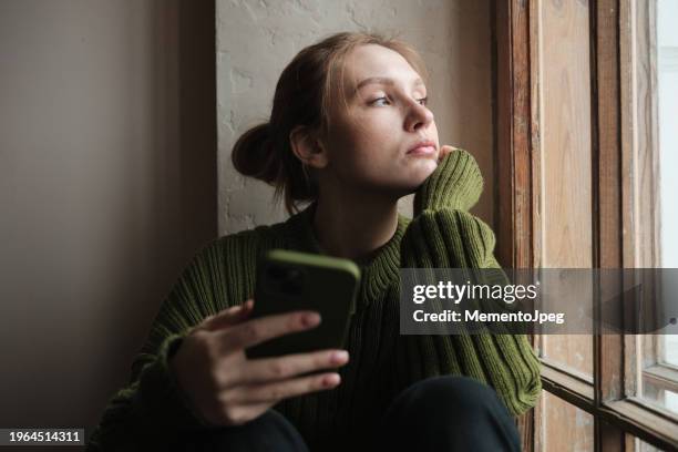 sad girl holding phone sitting on windowsill looking out window feeling down. social media and depression - problemas de parejas fotografías e imágenes de stock