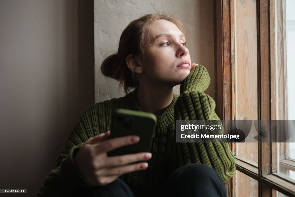 Sad girl holding phone sitting on windowsill looking out window feeling down. Social media and depression