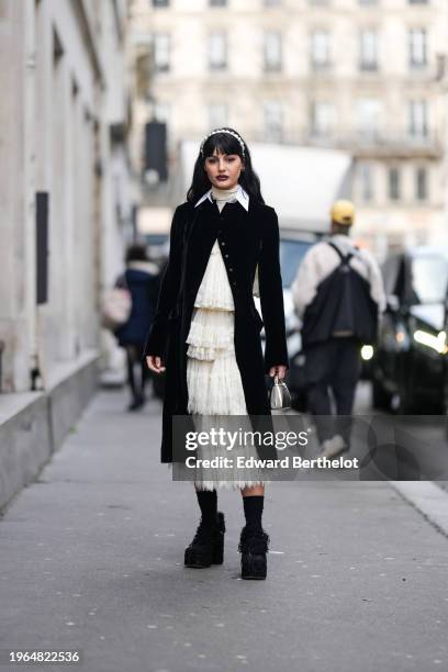 Maria Bernad wears a white headband, dark purple and black lipstick, make-up, earrings, a white turtleneck top, a black long jacket ,a white gathered...