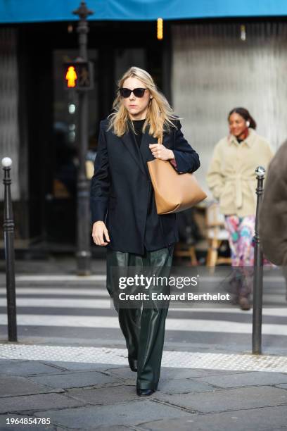 Guest wears sunglasses, a black oversized blazer jacket, a black top, a brown leather bag, green leather pants, leather shoes, outside Jean Paul...