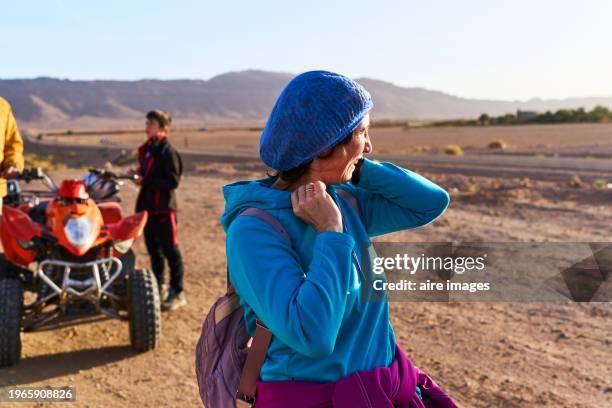 portrait of a female tourist in casual clothes standing in the desert of morocco looking away with motorcycle in the background, front view - trail moto maroc foto e immagini stock