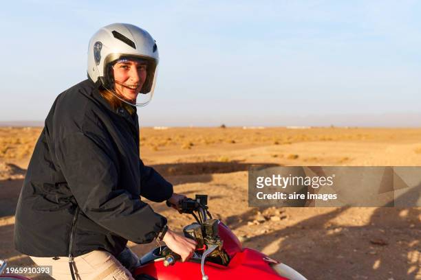 portrait of young woman wearing helmet sitting on four wheel motorbike smiling looking at camera, side view - trail moto maroc fotografías e imágenes de stock