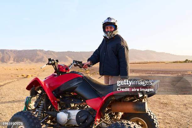 portrait of woman with helmet standing in morocco desert with four wheel motorbike looking at camera, front view - trail moto maroc foto e immagini stock