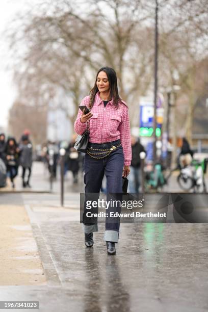 Guest wears a pink and white houndstooth pattern printed jacket, a black leather Chanel bag, a chain belt, blue cuffed jeans, black pointed boots,...