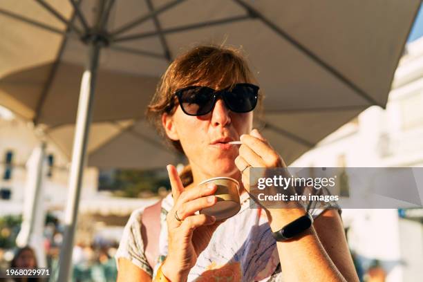 a woman seen from the front, wearing dark glasses, holding a cardboard cup, with a toothpick in her mouth, wearing a watch and rings on both hands, illuminated by the sun, the background is blurred. - com sombra imagens e fotografias de stock