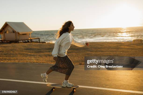 serious and concentrated woman honing her skateboard skills. riding at sunset by seaside - woman longboard stock-fotos und bilder