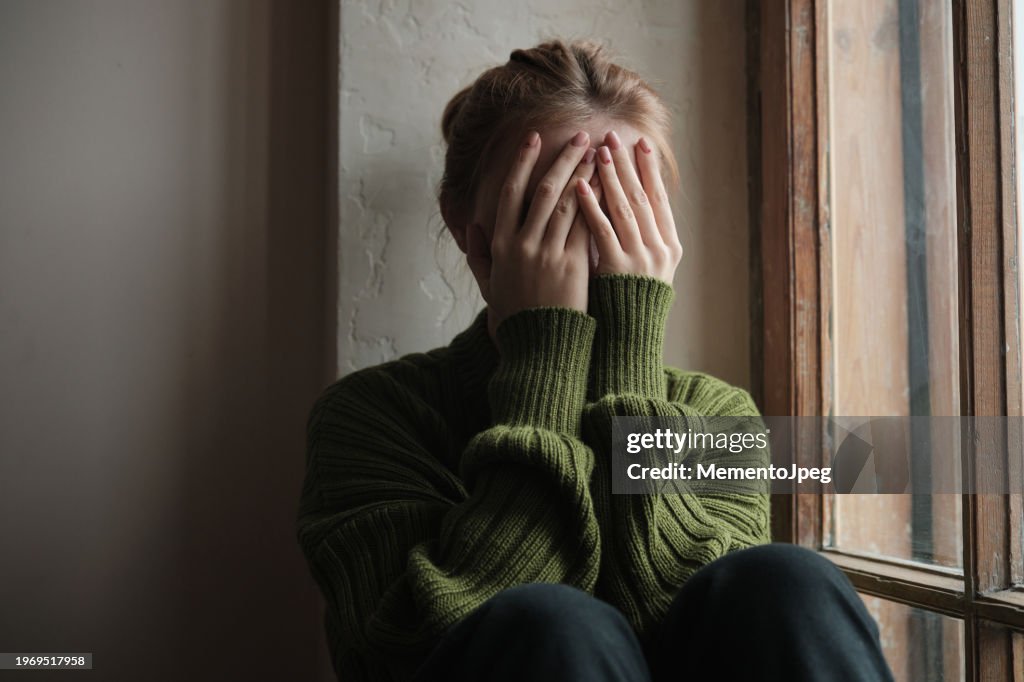 Upset depressed girl covering face with hands and crying while sitting on windowsill, sad worried teenager sitting by window, having problems. Teenagers and mental health concept